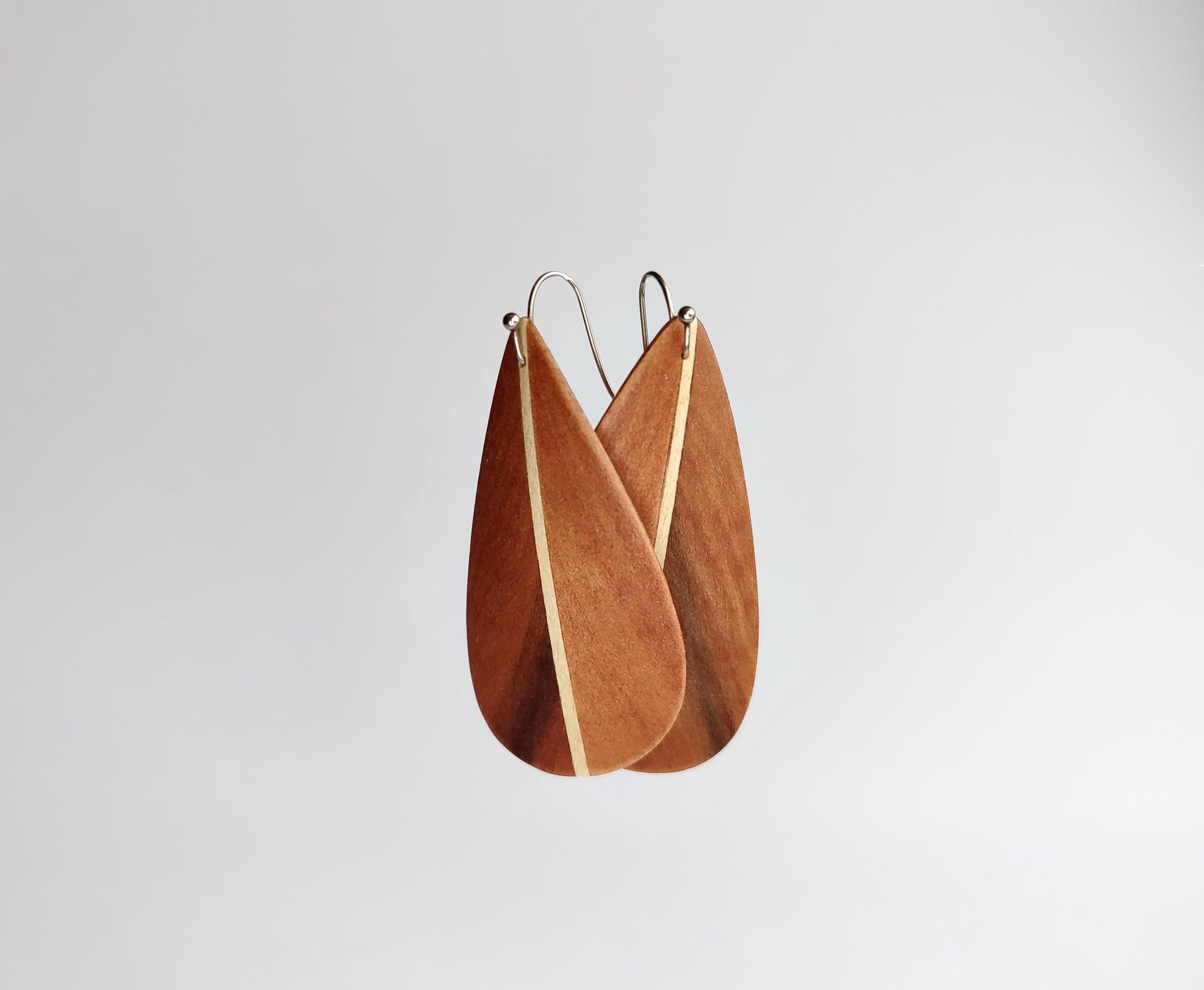 A pair of teardrop-shaped  feather inspired design wooden earrings with a natural wood grain finish and thin silver wire hooks, shown against a plain light gray background.