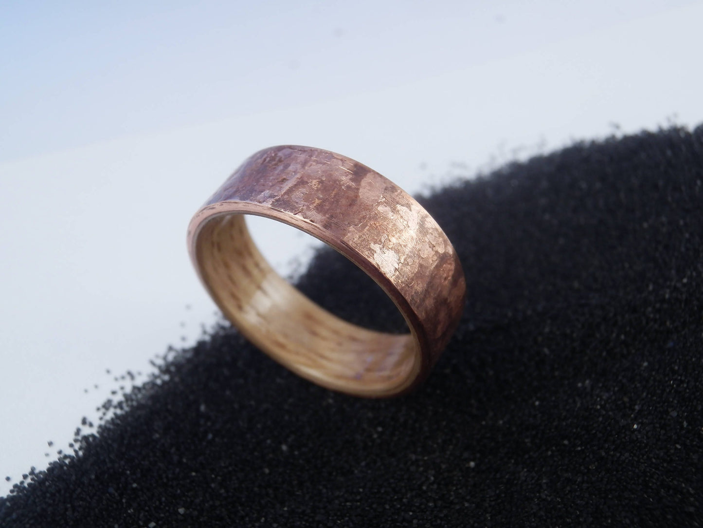 A hammered copper ring band resting on a bed of black sand against a light gray background.