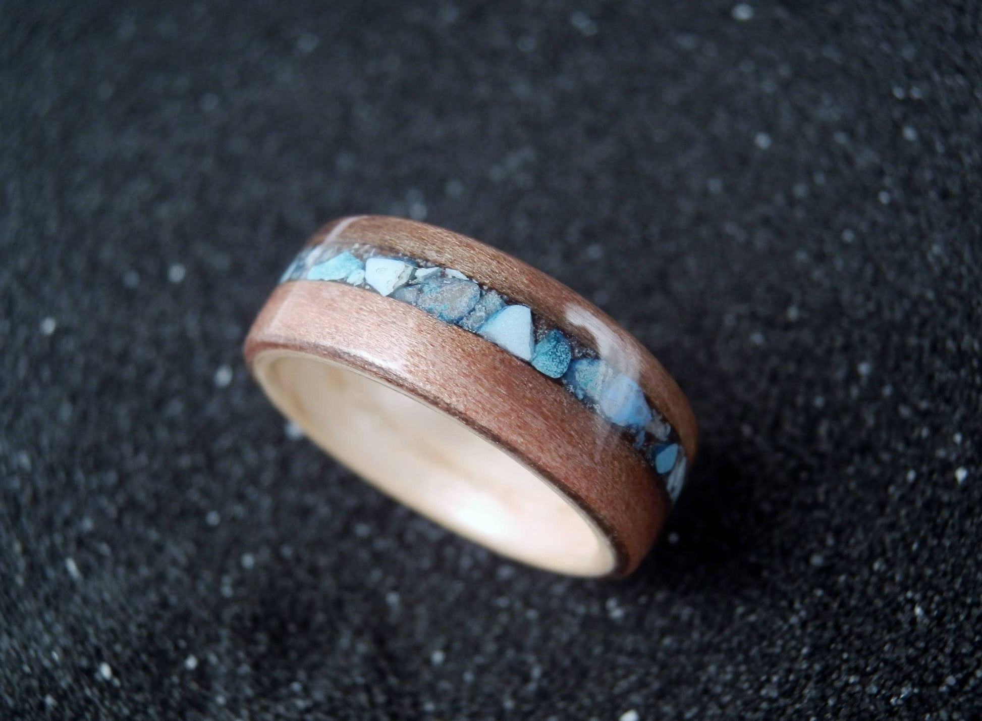 A wooden ring with a natural wood outer band, a central inlay of light blue and white marbled material, and a polished light wood inner band, resting on a dark textured surface.