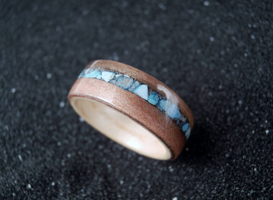 A wooden ring with a natural wood outer band, a central inlay of light blue and white marbled material, and a polished light wood inner band, resting on a dark textured surface.