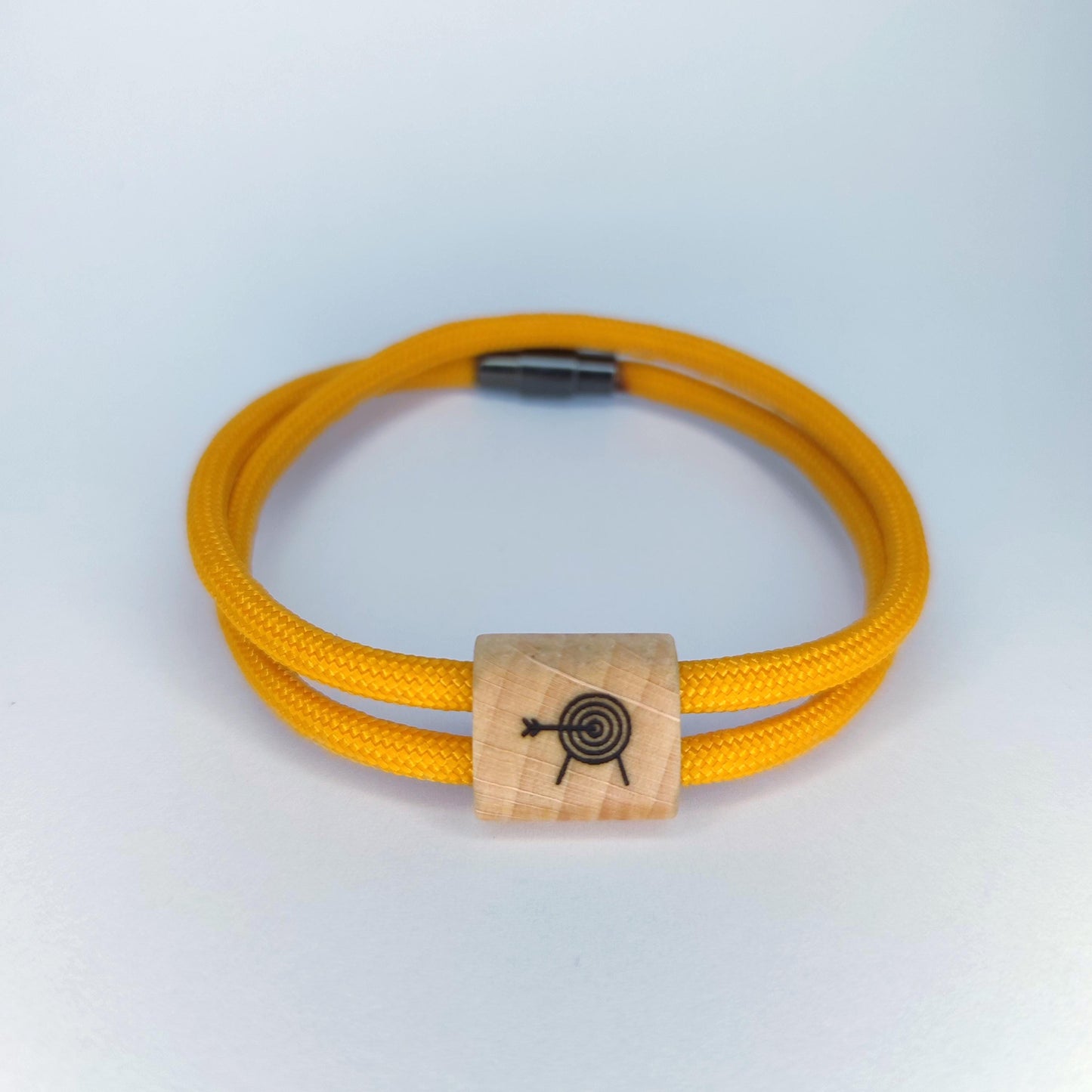 A yellow paracord bracelet with a rectangular light wood bead featuring a black engraved  archery target design, shown on a light gray background.
