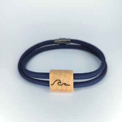 A double-strand navy blue braided cord bracelet with a natural light wood square bead engraved with a black wave design, shown on a light gray background.