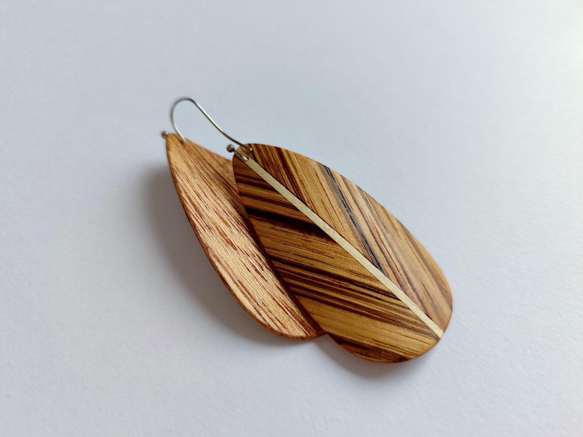 A pair of wooden dangle earrings shaped like elongated leaves, featuring natural wood grain patterns in shades of brown and beige, suspended from thin silver fishhook wires on a plain light background.