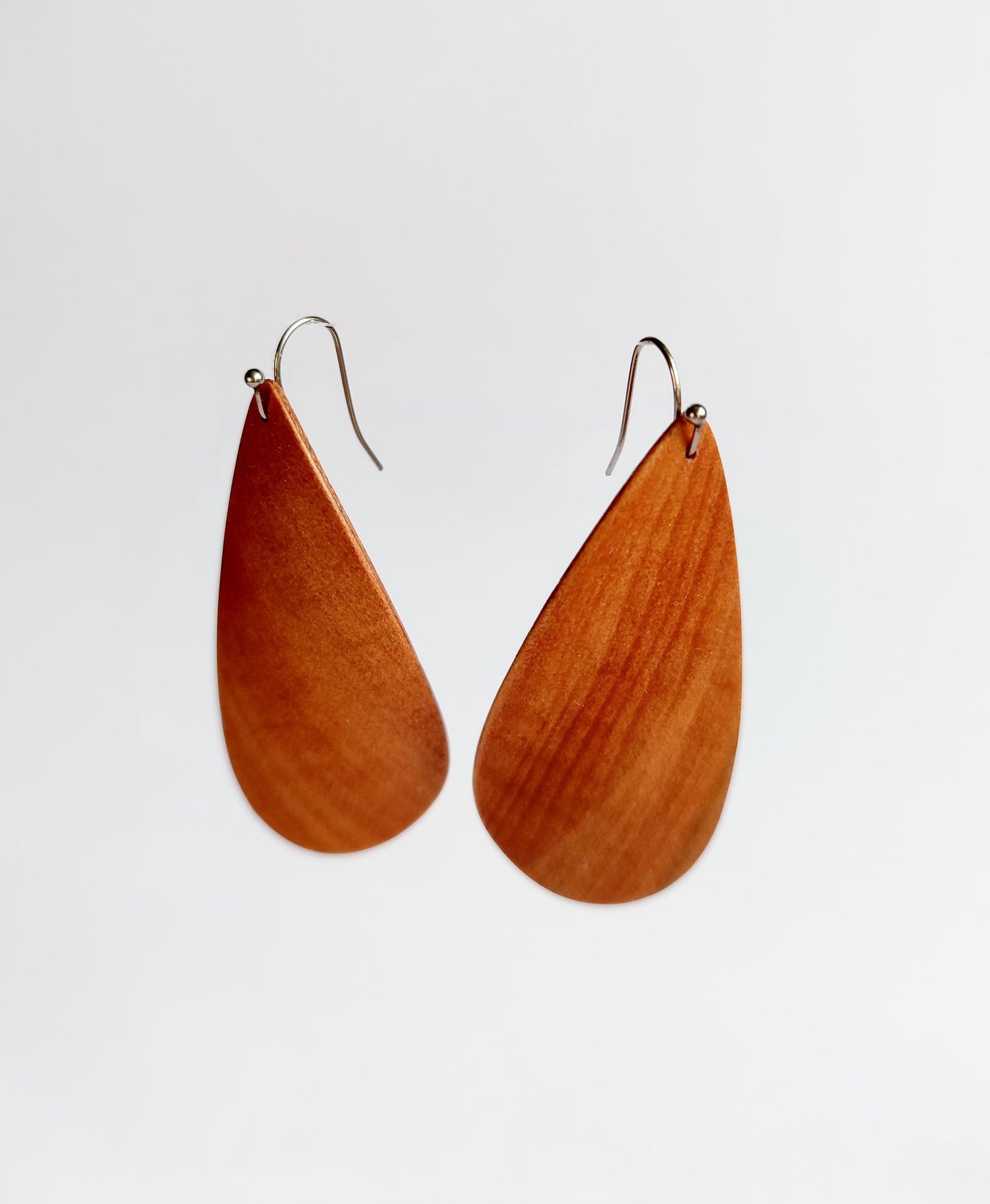 A pair of teardrop-shaped wooden drop earrings with a warm reddish-brown wood grain finish and silver-tone fishhook wires, shown against a plain light gray background.