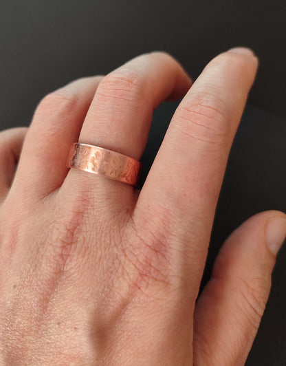 A close-up of a hand wearing a hammered copper band ring against a dark gray background.