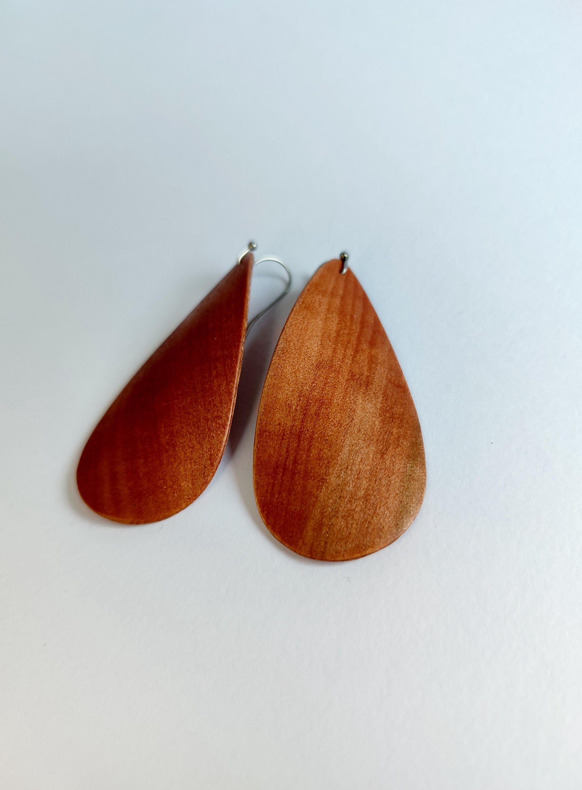A pair of teardrop-shaped wooden dangle earrings with a warm reddish-brown wood grain finish and silver-tone hooks, displayed on a plain off-white background.