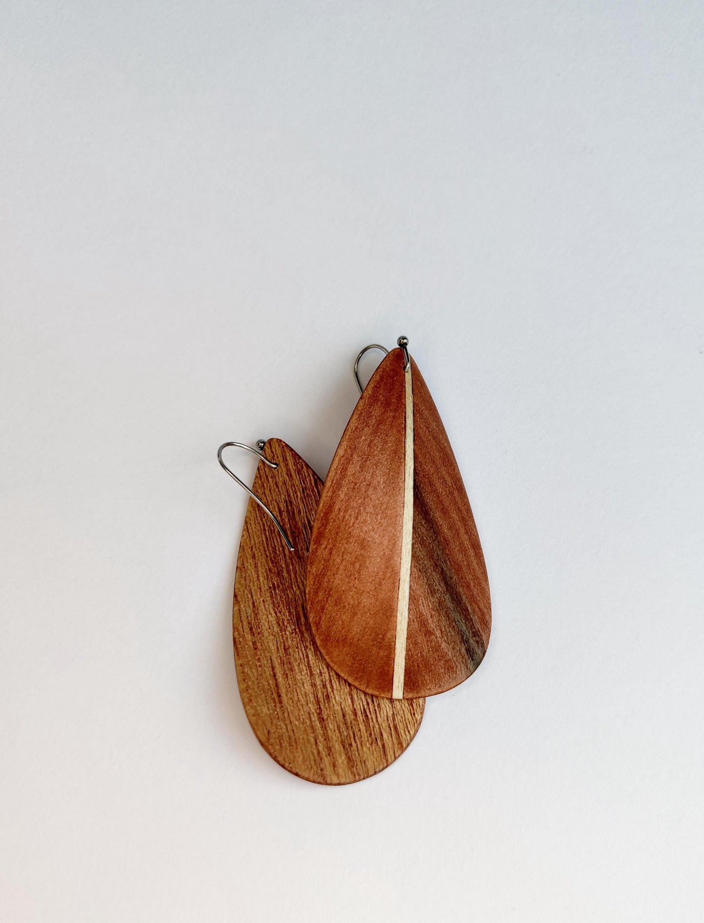 Teardrop-shaped wooden earrings with a warm reddish-brown wood grain and a thin light beige inlay, hanging from silver fishhook wires against a plain white background.