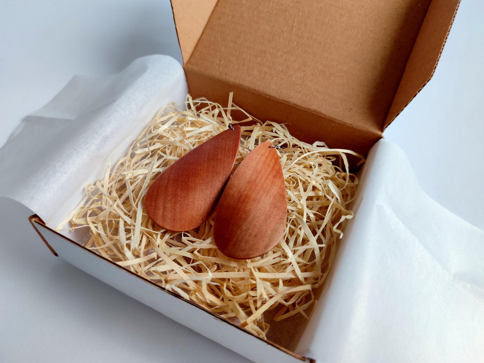 Two small teardrop-shaped reddish-brown wooden pieces placed in shredded paper inside an open brown cardboard box lined with white tissue paper, on a light gray background.