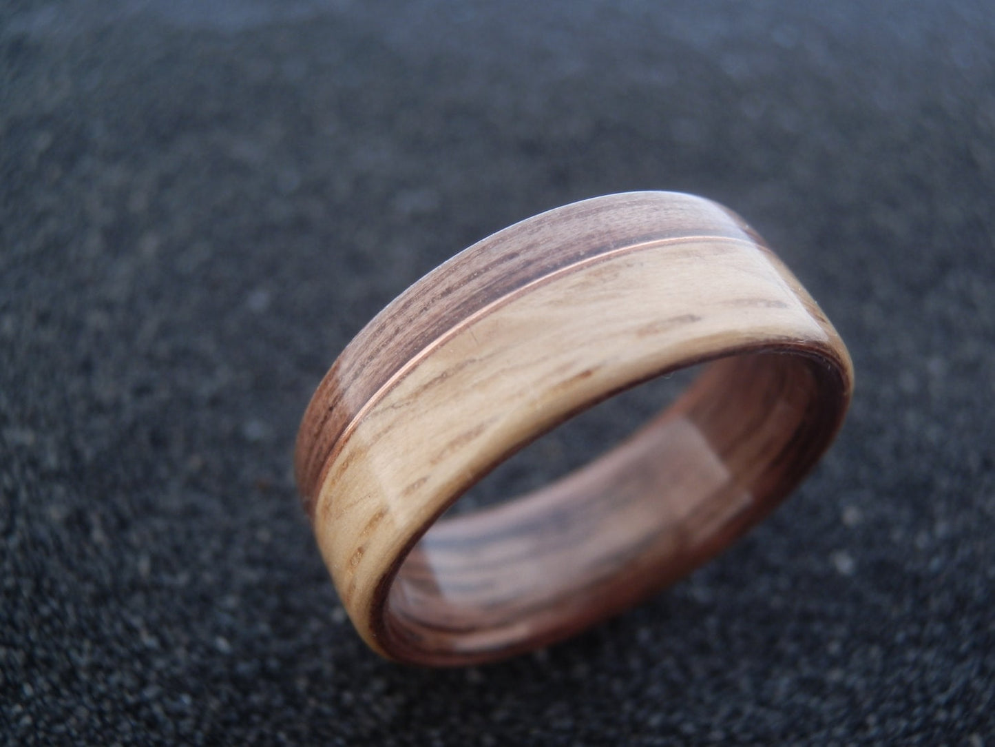 A wooden ring band with a natural wood grain pattern in light tan and dark brown, resting on a dark textured surface.