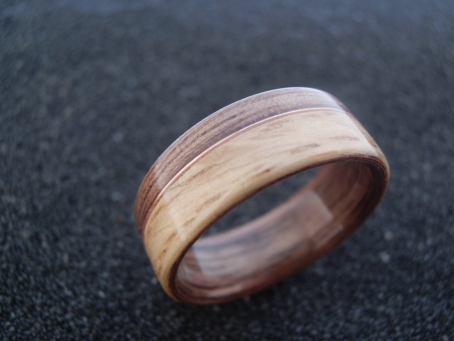 A wooden ring band with a natural wood grain pattern in light tan and dark brown, resting on a dark textured surface.