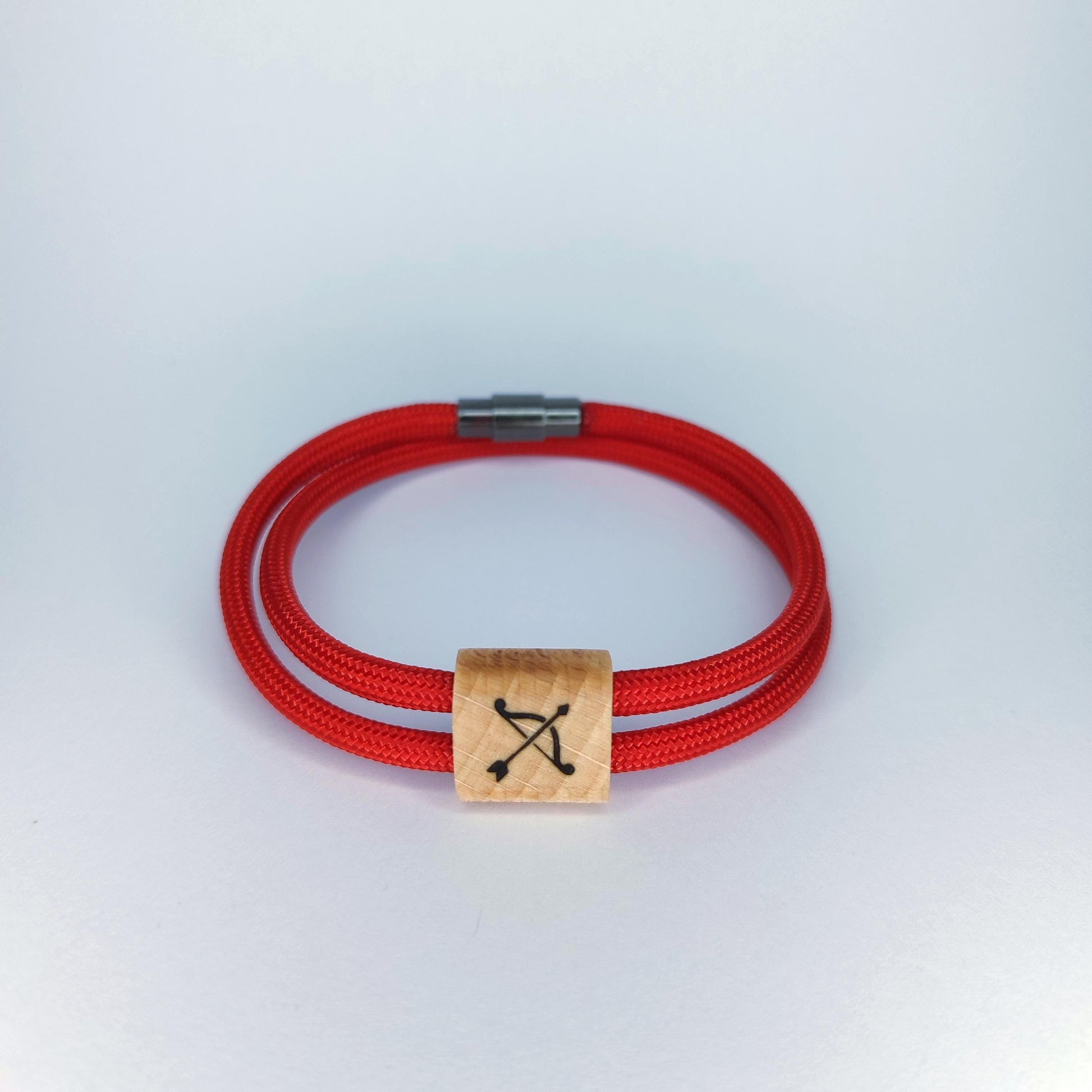 A red paracord bracelet with a square light wood charm engraved with a black bow and arrow symbol, resting on a light gray background.