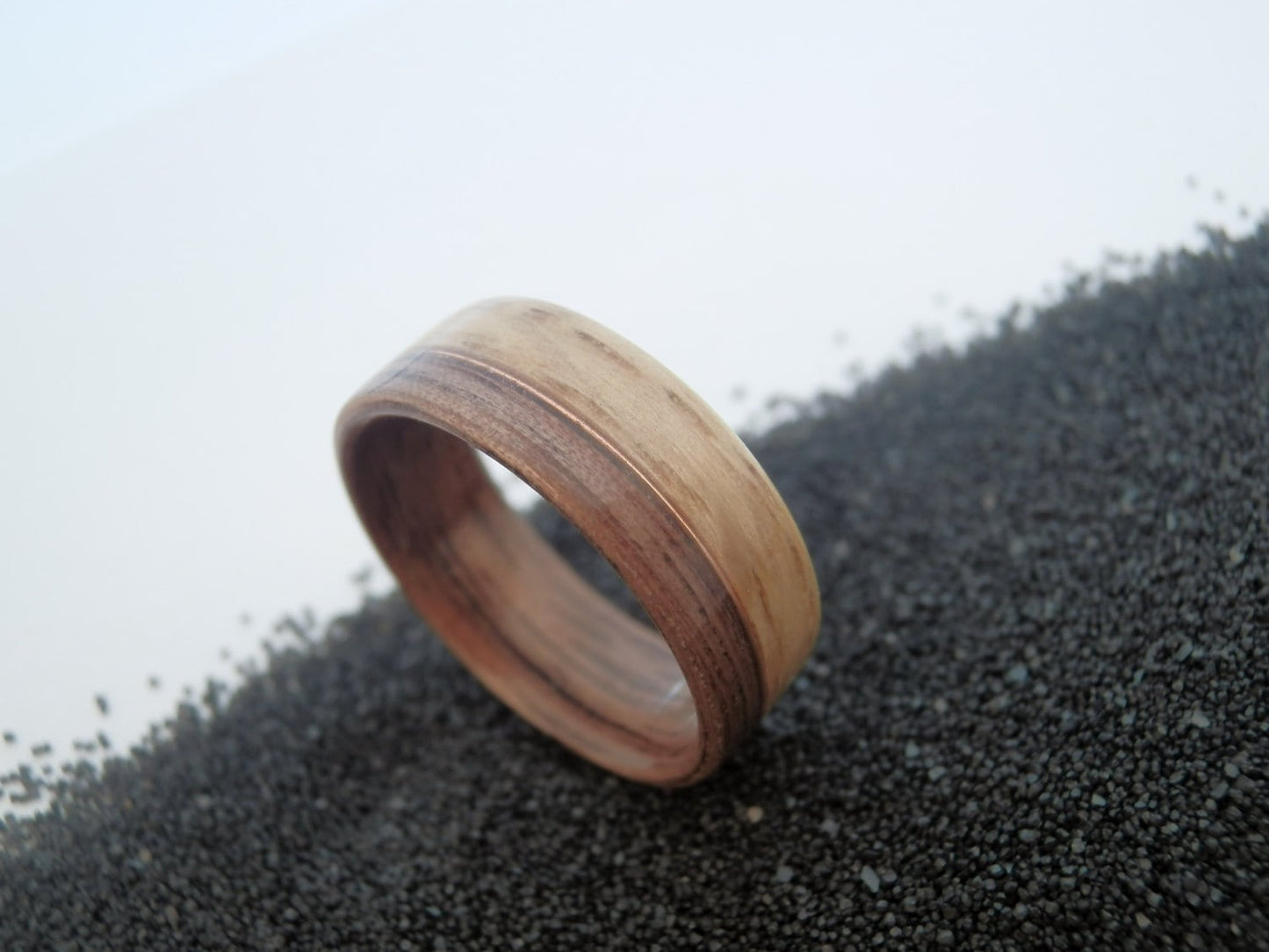 Walnut and oak ring band with a natural wood grain pattern, resting on a bed of small dark gravel against a light gray background.