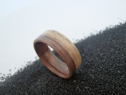 Walnut and oak ring band with a natural wood grain pattern, resting on a bed of small dark gravel against a light gray background.