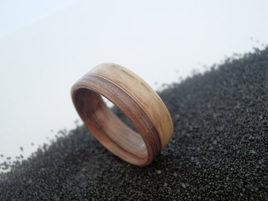 Walnut and oak ring band with a natural wood grain pattern, resting on a bed of small dark gravel against a light gray background.