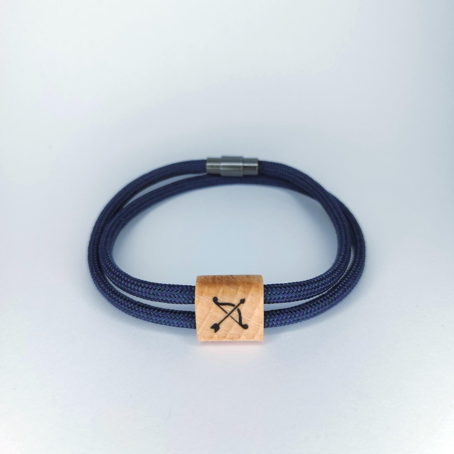 A double-strand navy blue braided cord bracelet with a square light wood charm engraved with a black bow and arrow symbol, finished with a silver metal clasp, shown on a light gray background.