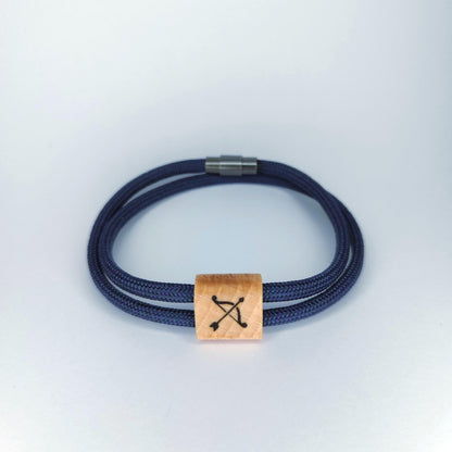 A double-strand navy blue braided cord bracelet with a square light wood charm engraved with a black bow and arrow symbol, finished with a silver metal clasp, shown on a light gray background.