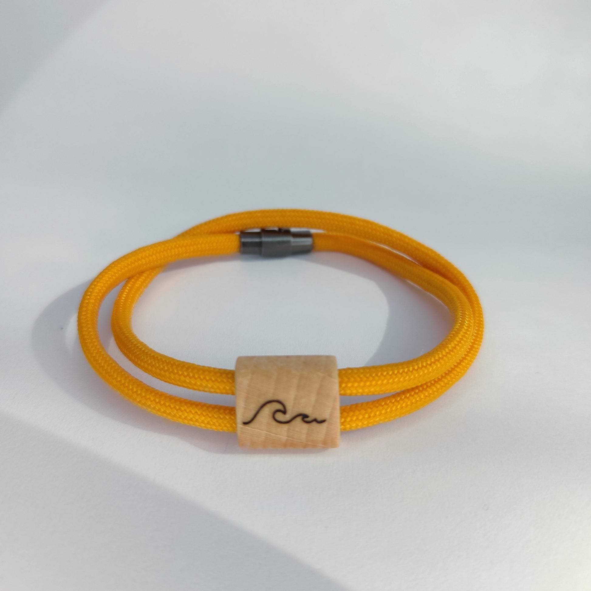 A yellow paracord bracelet with a natural wood square charm engraved with a black wave, secured by a silver metal clasp, on a white background.