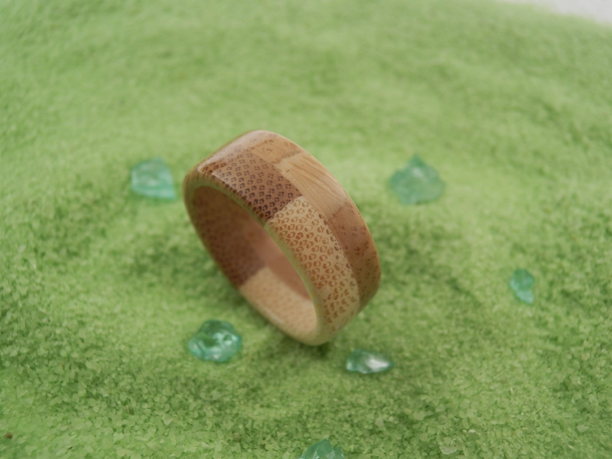 A handmade bamboo ring with a checkerboard pattern of light and dark wood grain, displayed on a textured green background with small scattered green stones.
