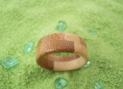 A handmade bamboo ring with a textured honeycomb pattern and natural wood grain, resting on a green textured surface with small decorative green stones.