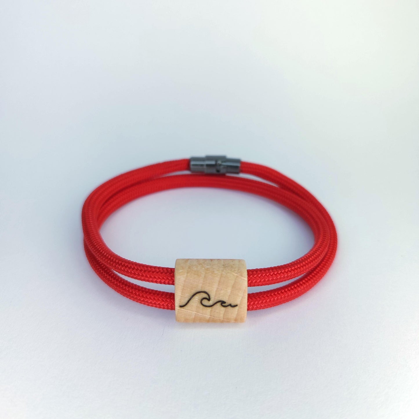 A red paracord bracelet with a natural wood square charm engraved with a black wave symbol, set against a plain white background.