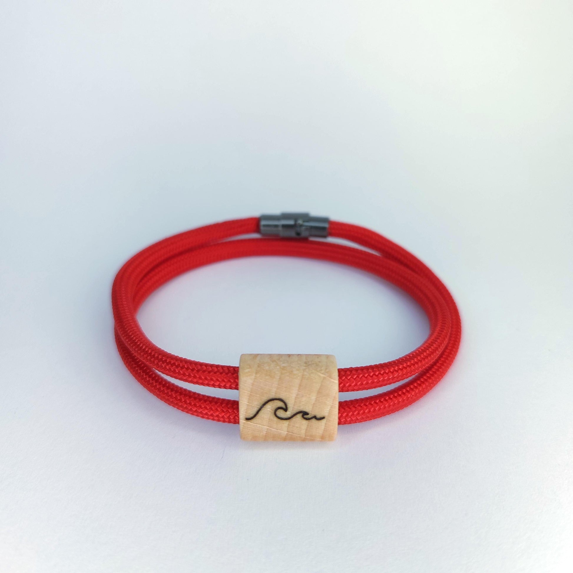 A red paracord bracelet with a natural wood square charm engraved with a black wave symbol, set against a plain white background.