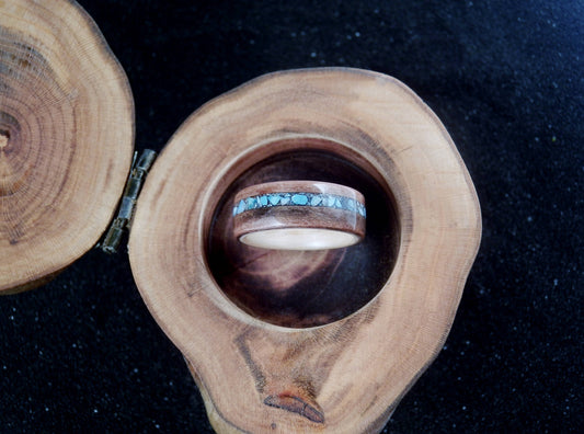 A wooden ring box with a natural wood slice lid and a circular wooden tray holding a wood ring with a turquoise inlay, set against a dark background.
