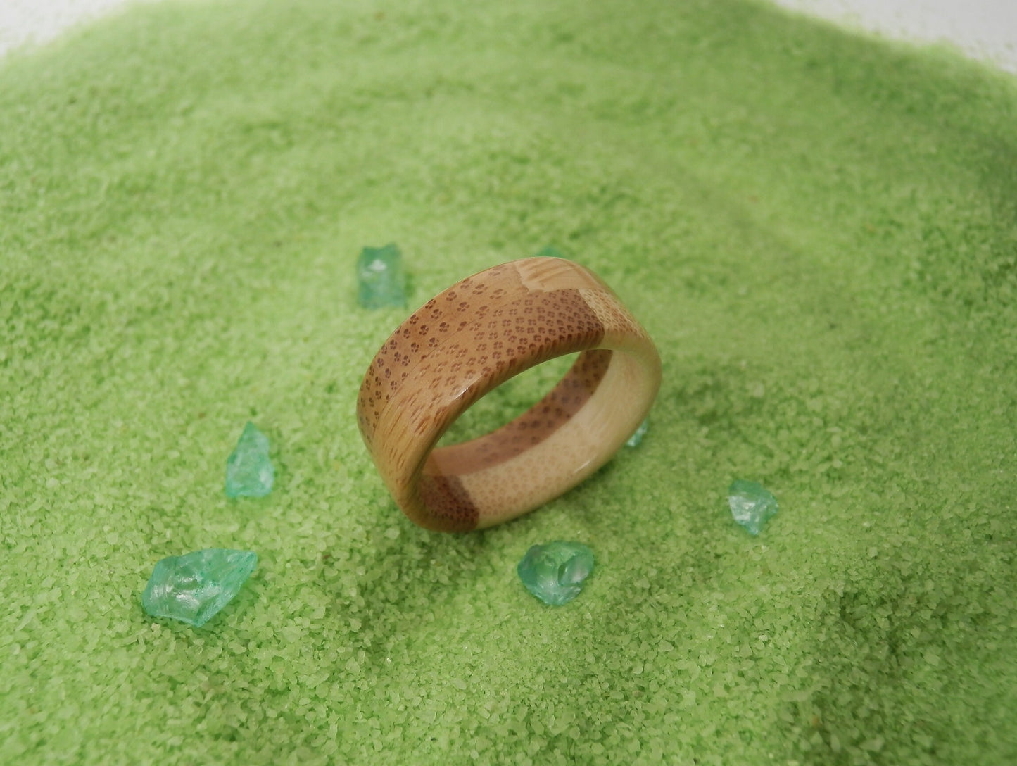 A textured bamboo ring with a honeycomb pattern and a polished brown center band, resting on a bed of green powder with small green crystal accents.