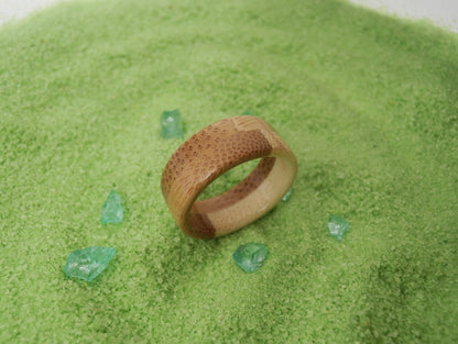 A textured bamboo ring with a honeycomb pattern and a polished brown center band, resting on a bed of green powder with small green crystal accents.