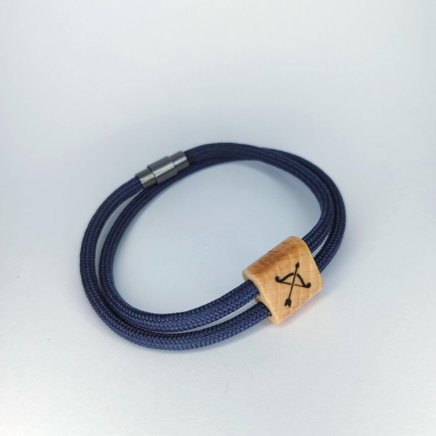 A navy blue braided cord bracelet with a silver metal clasp and a light wood square charm engraved with an arrow motif, shown on a light gray background.