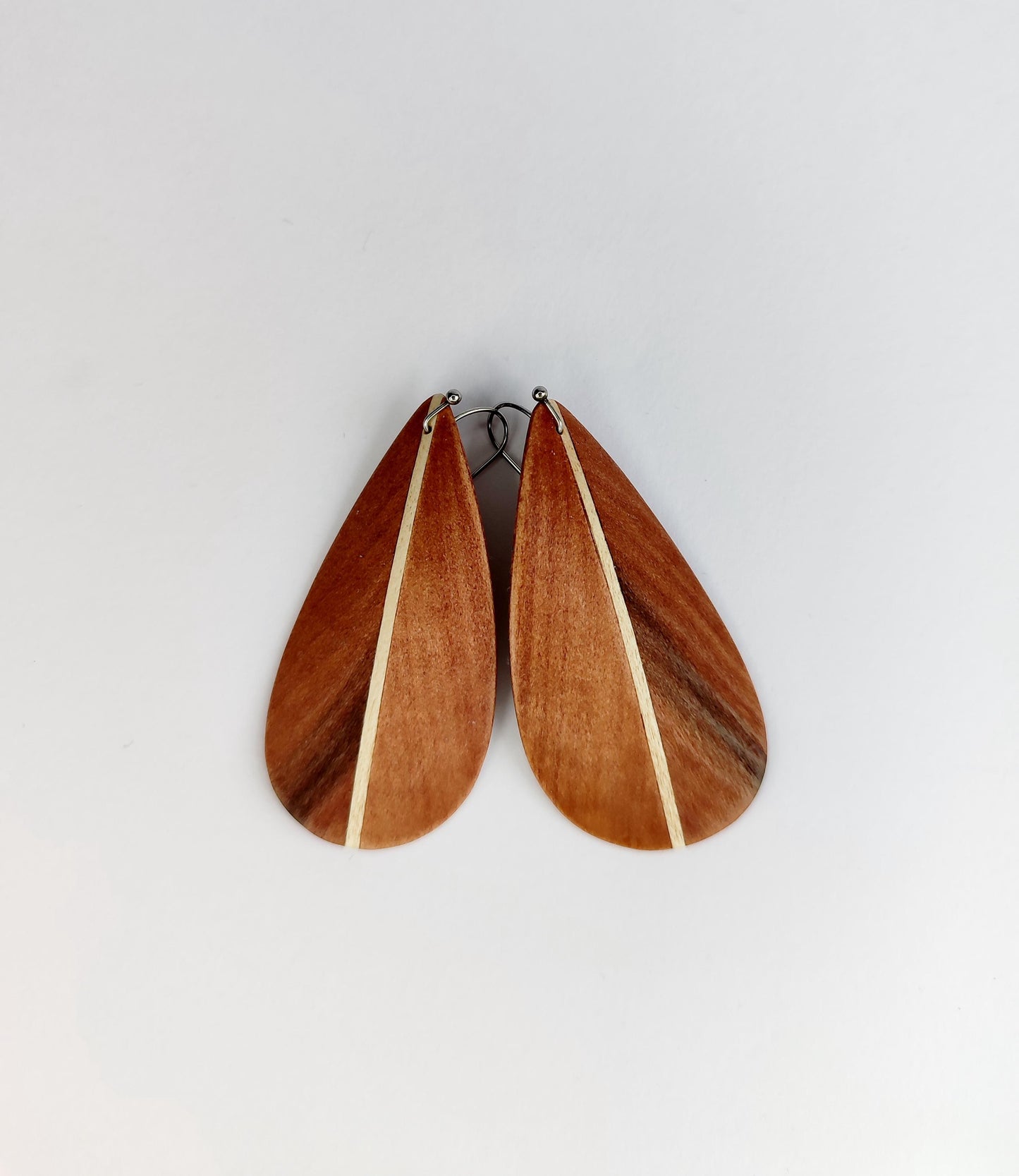 A pair of teardrop-shaped wooden earrings with a warm reddish-brown wood grain and a thin light beige inlay, displayed against a plain light gray background.