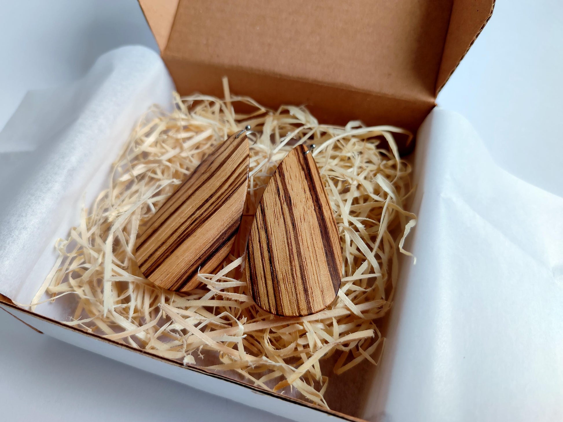 A pair of teardrop-shaped wooden earrings with dark grain patterns, nestled in shredded paper inside an open brown cardboard gift box.