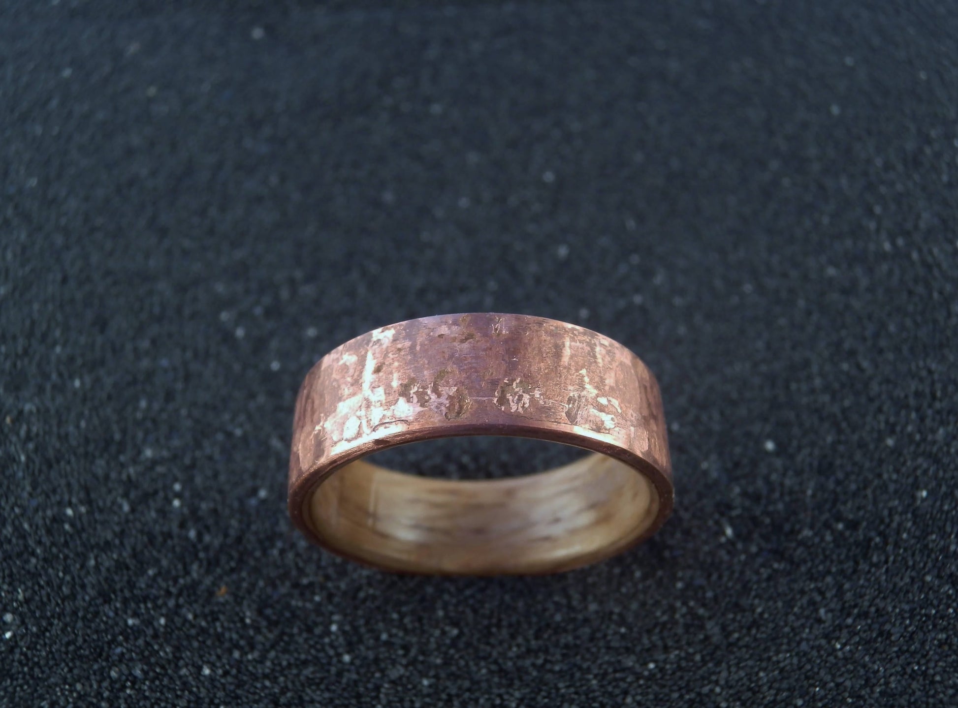 A hammered copper and oak wood band ring with a textured, metallic finish, displayed on a dark, textured background.