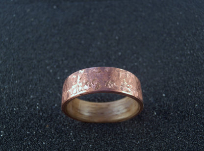 A hammered copper and oak wood band ring with a textured, metallic finish, displayed on a dark, textured background.